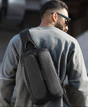 Man wearing sunglasses and a gray sweatshirt, carrying a black BANGE tech sling bag outdoors.