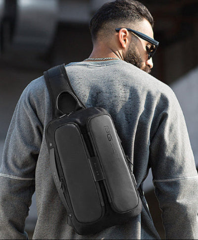Man wearing sunglasses and a gray sweatshirt, carrying a black BANGE tech sling bag outdoors.