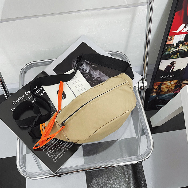 Beige crossbody sling bag with bright orange zipper pulls, displayed on a transparent chair for a modern aesthetic.