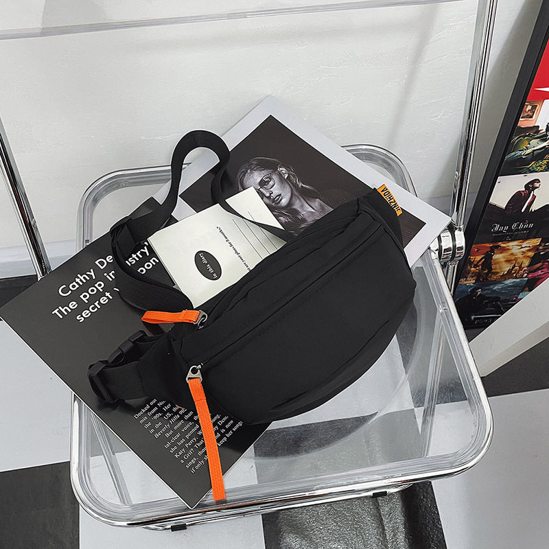 Black crossbody sling bag with orange zipper accents, placed on a clear acrylic chair with lifestyle books.
