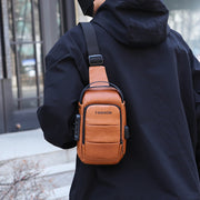 Brown men’s leather sling bag with USB charging port worn over the shoulder outdoors.