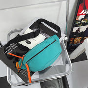 Teal crossbody sling bag with bold orange zipper pulls, styled on a clear chair with monochrome magazines.