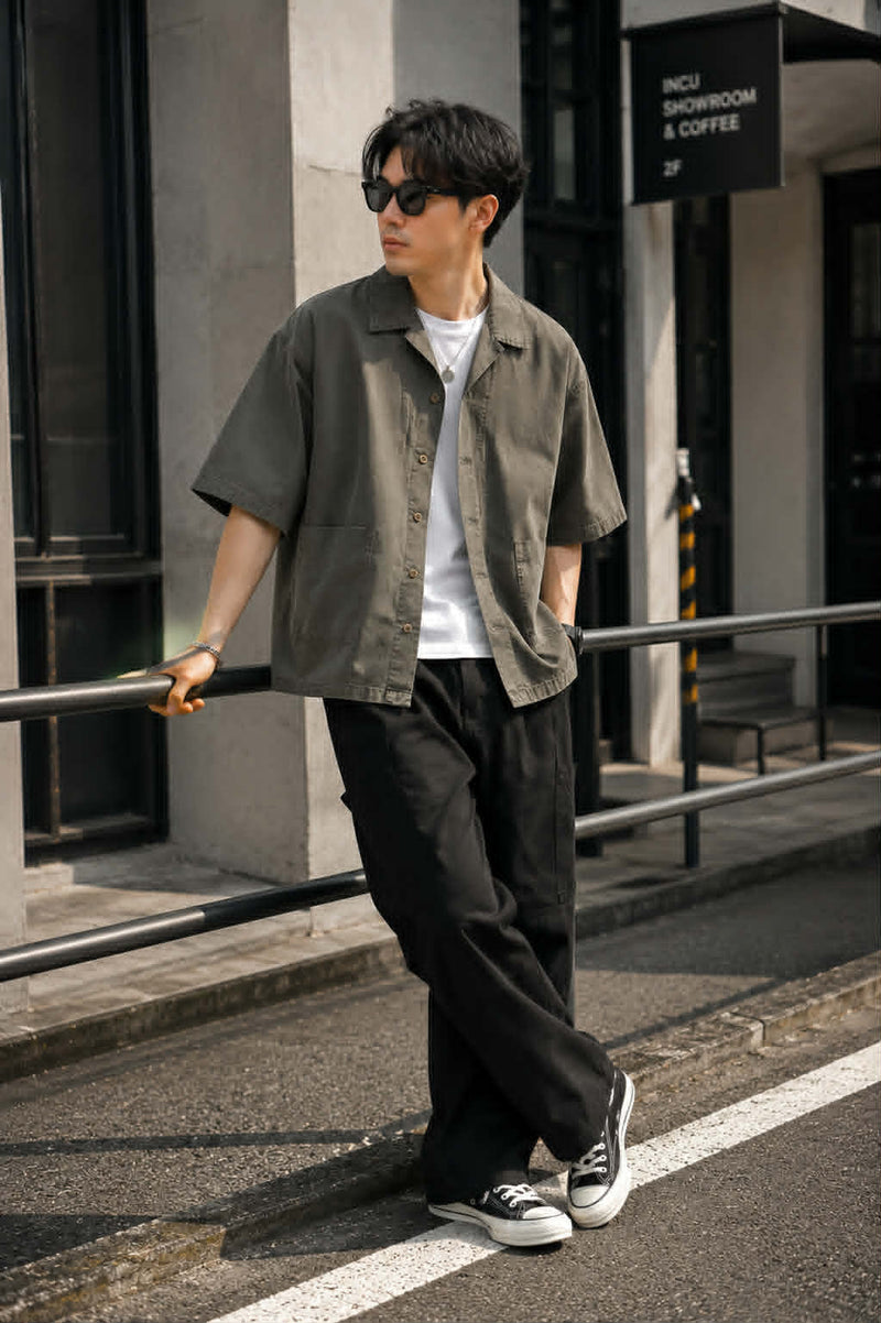 Male model wearing an oversized dark gray Japanese workwear Cuban collar shirt with rounded patch pockets, styled with a white t-shirt, black cargo pants, and sneakers in an urban street setting.
