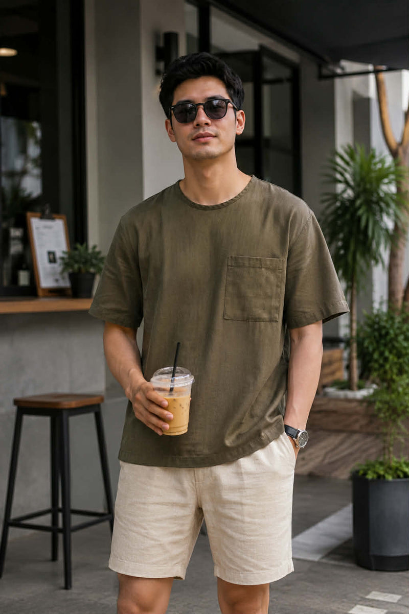 Men's olive green relaxed fit cotton pocket t-shirt styled with beige shorts in a modern cafe setting
