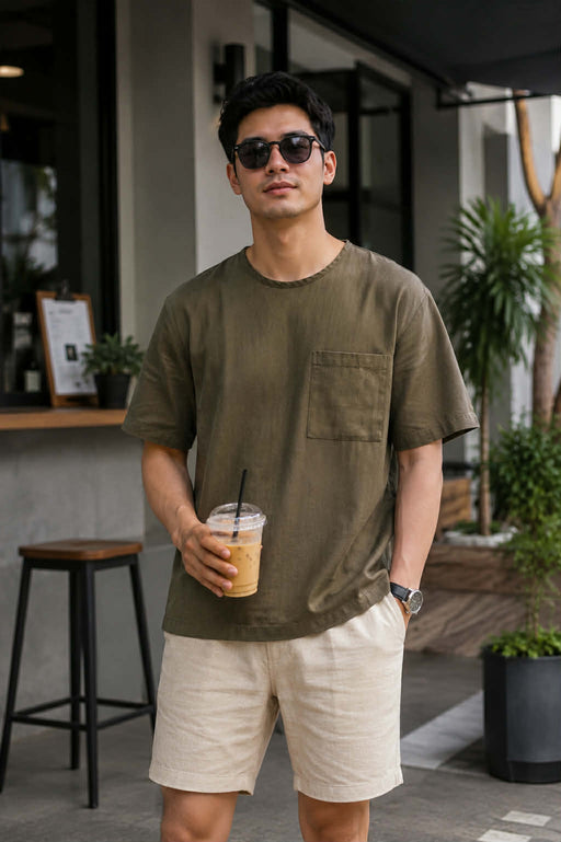 Men's olive green relaxed fit cotton pocket t-shirt styled with beige shorts in a modern cafe setting
