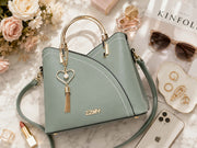 Luxury flatlay featuring a sage green structured handbag with gold hardware and heart pearl tassel charm, surrounded by perfume, sunglasses, jewelry, flowers, lipstick, and smartphone on a marble tabletop.
