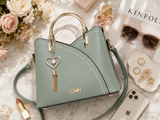 Luxury flatlay featuring a sage green structured handbag with gold hardware and heart pearl tassel charm, surrounded by perfume, sunglasses, jewelry, flowers, lipstick, and smartphone on a marble tabletop.
