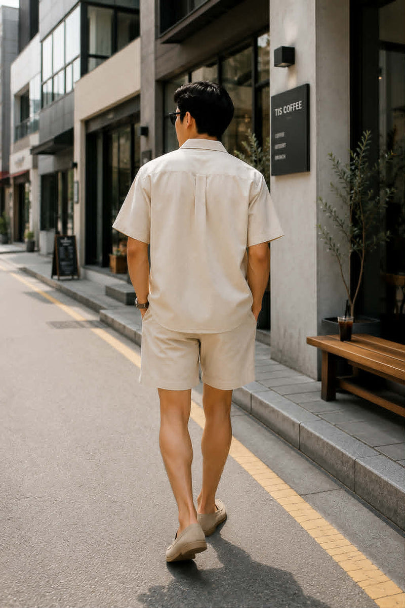 Back view of an Asian male model wearing a men's beige retro loose fit short sleeve button-up shirt and beige shorts walking on a modern city street.
