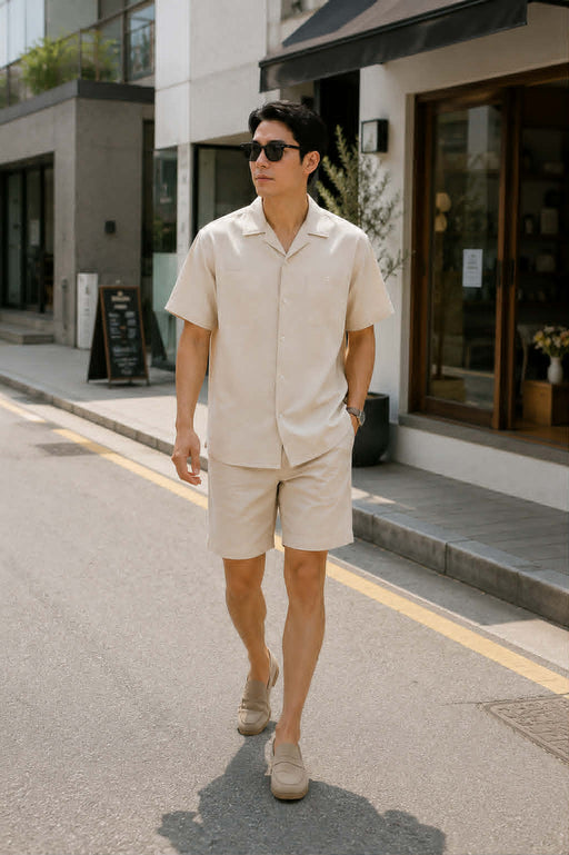 Asian male model wearing a men's beige retro loose fit short sleeve button-up shirt with matching shorts and white sneakers walking on a modern urban street.
