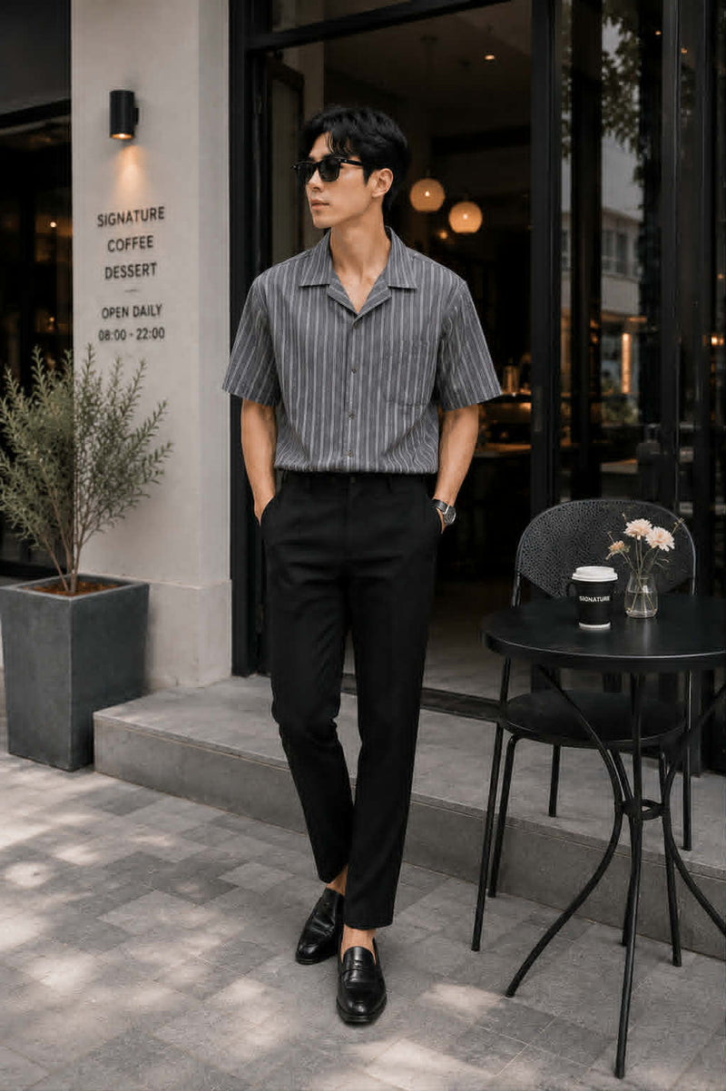 Asian male model wearing a gray retro Cuban collar striped short sleeve shirt tucked into black tailored trousers with leather loafers, showcasing a sophisticated smart casual Korean fashion look outside a luxury café.

