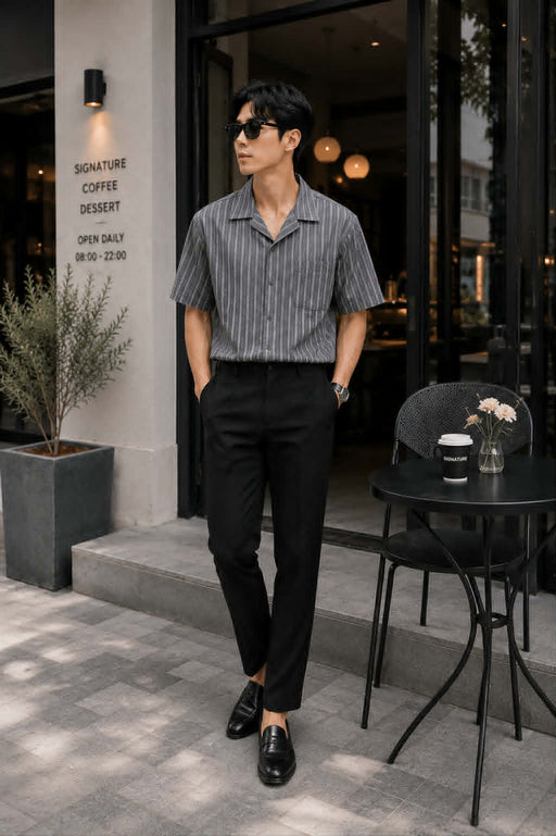 Asian male model wearing a gray retro Cuban collar striped short sleeve shirt tucked into black tailored trousers with leather loafers, showcasing a sophisticated smart casual Korean fashion look outside a luxury café.

