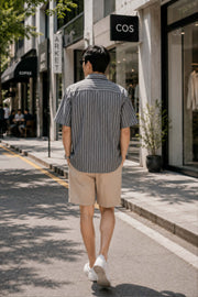 Back view of an Asian male model wearing a gray retro Cuban collar striped short sleeve shirt with beige shorts and white sneakers, walking through a modern shopping district in a relaxed Korean streetwear style.
