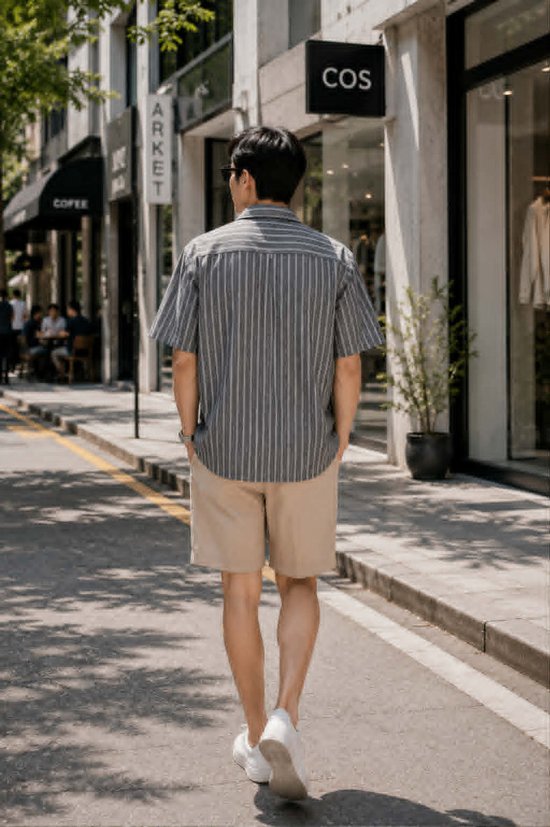 Back view of an Asian male model wearing a gray retro Cuban collar striped short sleeve shirt with beige shorts and white sneakers, walking through a modern shopping district in a relaxed Korean streetwear style.
