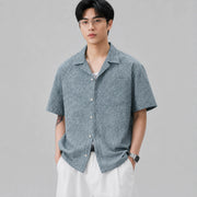 Front view of men's textured blue Cuban collar short sleeve shirt styled with white pants.
