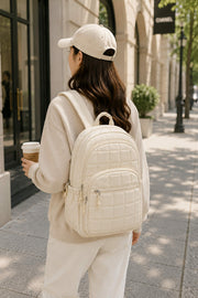 Woman wearing an off white quilted backpack while walking in the city holding a coffee, showcasing a stylish and lightweight everyday travel backpack.

