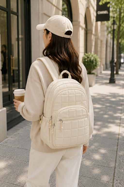 Woman wearing an off white quilted backpack while walking in the city holding a coffee, showcasing a stylish and lightweight everyday travel backpack.
