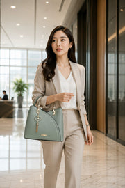 Elegant professional woman carrying a sage green structured handbag with gold metal handles and heart tassel charm while walking through a luxury shopping district, wearing a sophisticated business casual outfit in natural daylight.
