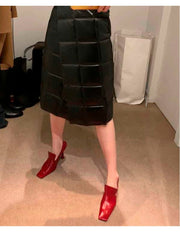 A person wearing a black quilted pleated midi skirt and bright burgundy pointed-toe heeled mules, standing on a beige carpeted floor in an indoor room.