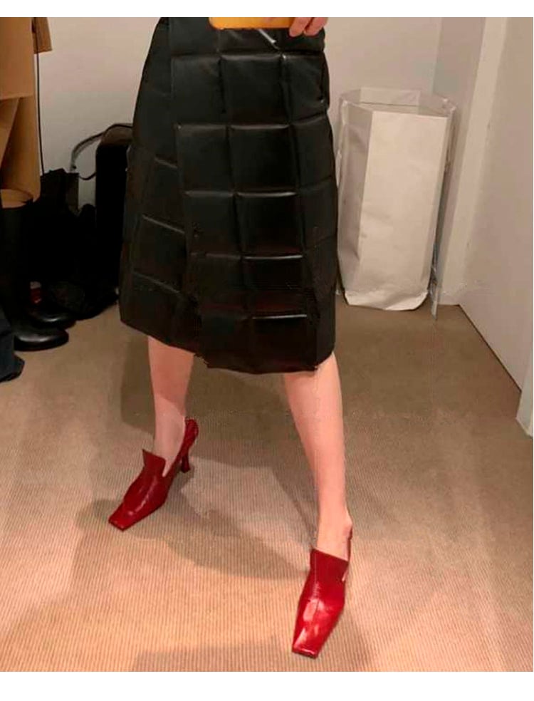 A person wearing a black quilted pleated midi skirt and bright burgundy pointed-toe heeled mules, standing on a beige carpeted floor in an indoor room.