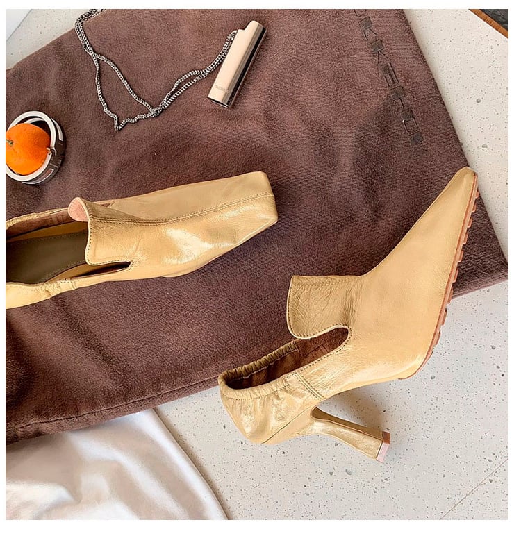 A pair of beige leather ankle boots with kitten heels, styled on a brown suede-like fabric background with a silver chain necklace and a small orange candle.
